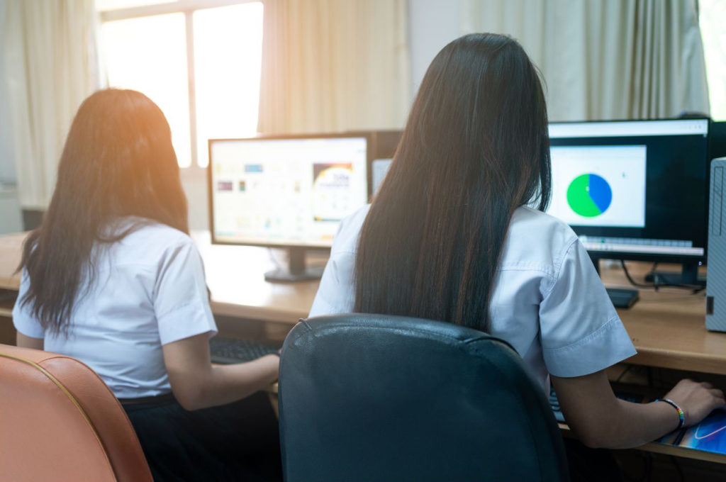 ２人の女子大生がデスクトップパソコンで調べ物をしている