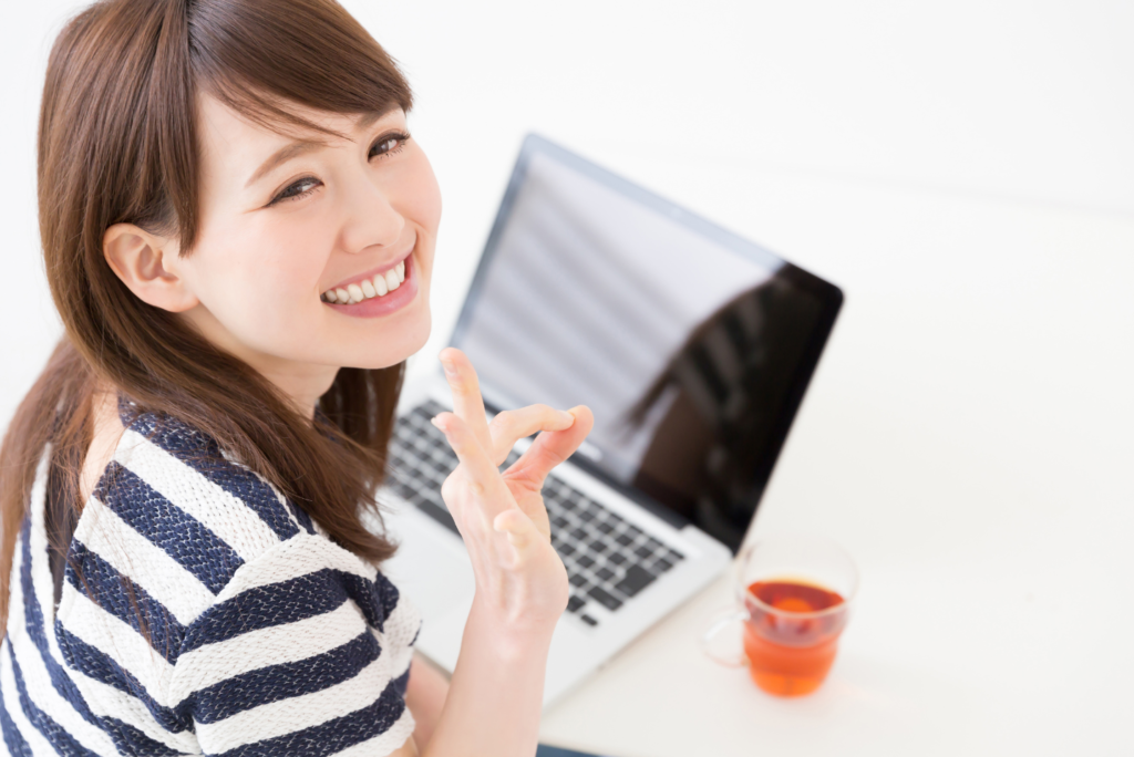 ノートパソコンをいじりながら振り向きざまに右手でOKサインをしながら微笑む若い女性